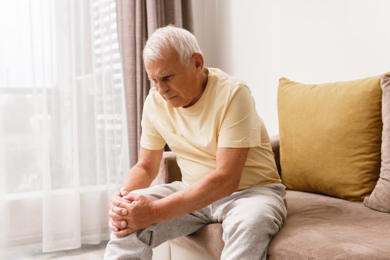 An elderly man sitting on the edge of a bed, holding his painful knee in discomfort after a busy day.
