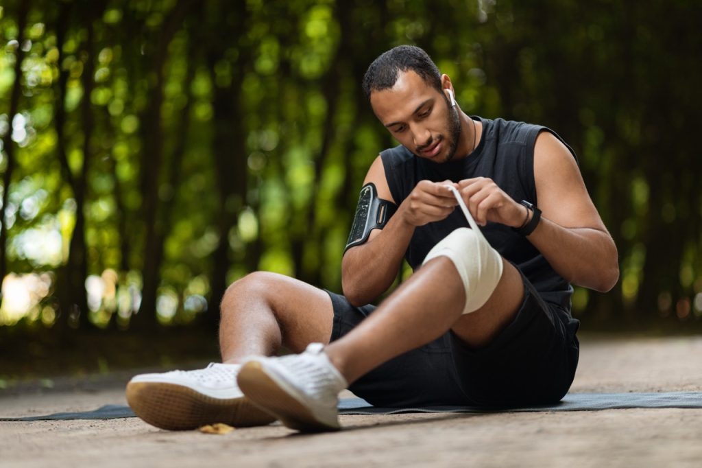 Athletic man wrapping an elastic bandage around his knee due to knee pain exercise.