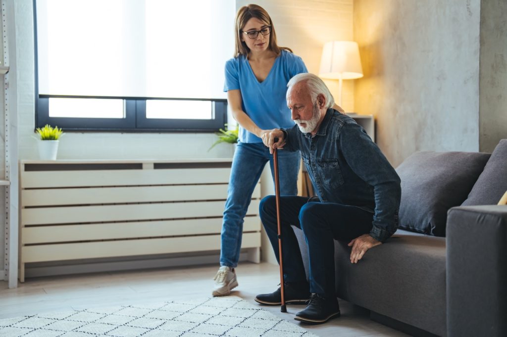 Physical therapist assisting an elderly man standing up from a sofa with a cane