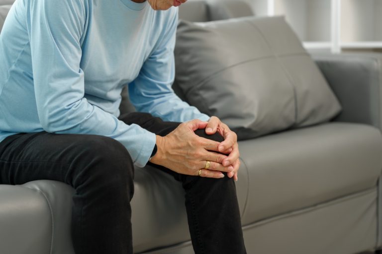 Older man sitting on a sofa clutching an inflamed knee in pain.