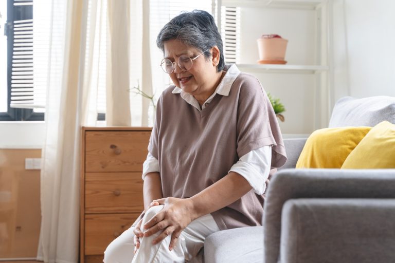 A close-up of a person sitting down and applying a clear topical pain relief gel from a white tube onto an inflamed, red knee joint.