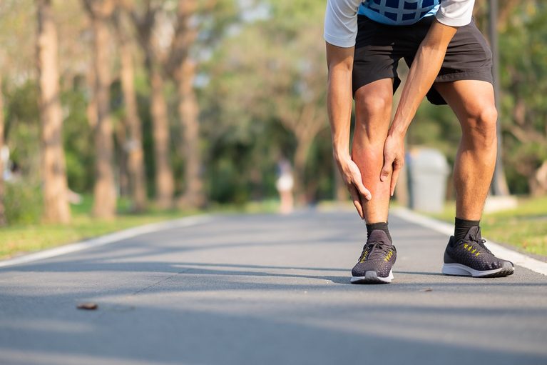 A man in black athletic wear bending over and holding his knee in pain while on an outdoor running track.
