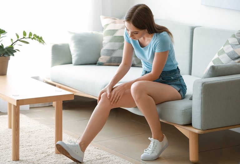 Young woman sitting on a couch and holding her painful knee at home