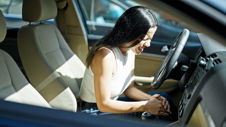 A woman sitting in the driver's seat of a car, holding her knee in pain while leaning forward.